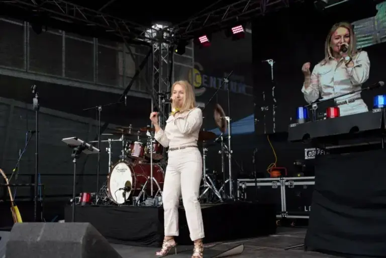 Corinna Anders in Weiß singt auf einer Freilichtbühne vor Schlagzeug und Leinwand