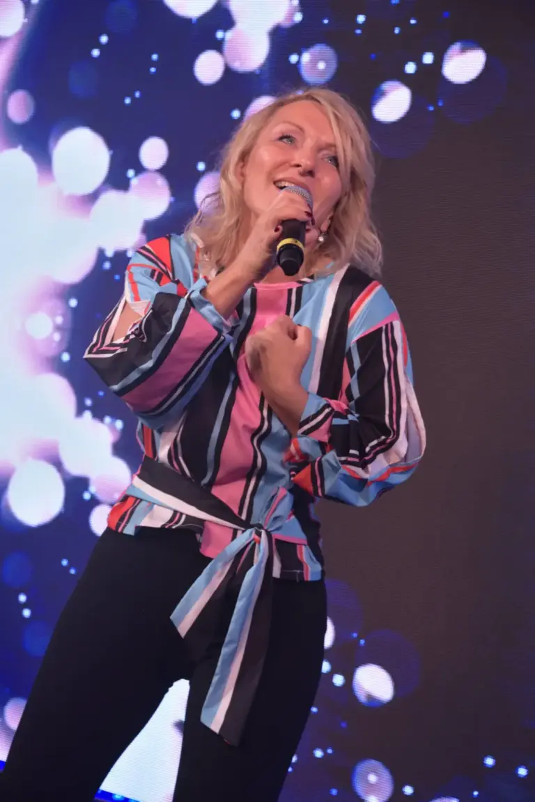 Corinna Anders singt in bunt gestreifter Bluse vor angestrahlter Kulisse