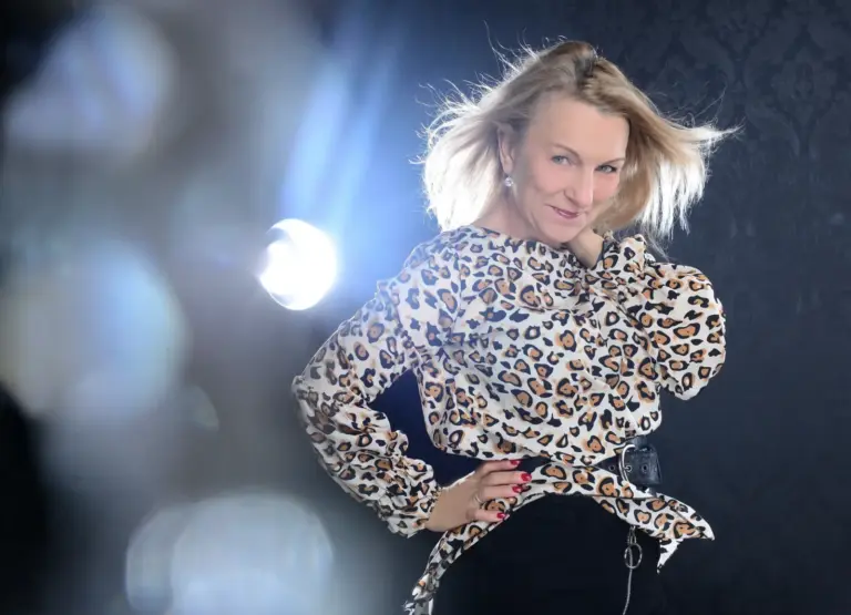 Corinna Anders in Leopardenbluse mit Händen in der Hüfte, Licht hinter ihr