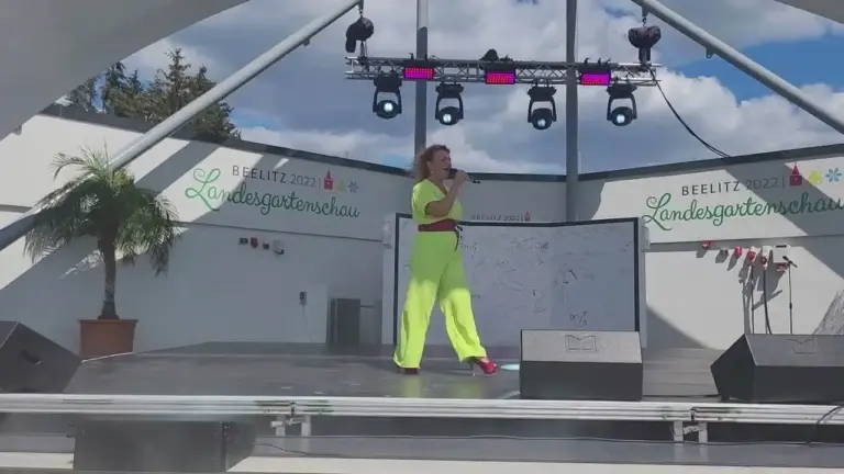 Corinna Anders in Neongelb singt auf einer Freilichtbühne mit Palme im Hintergrund