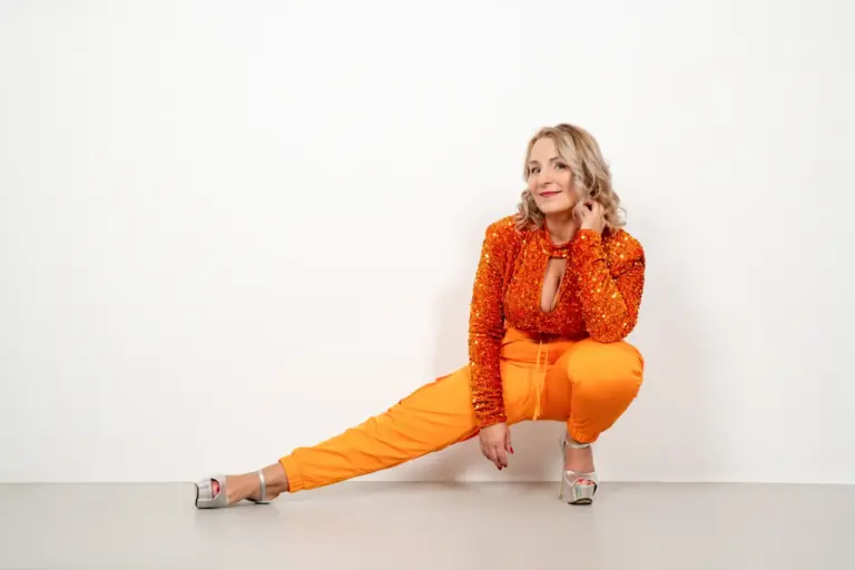 Corinna Anders im orangefarbenen Paillettentop mit seitlich ausgestrecktem Bein