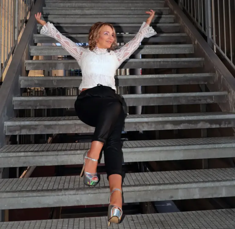 Corinna Anders sitzt im weißen Spitzentop auf einer Treppe, die Arme erhoben